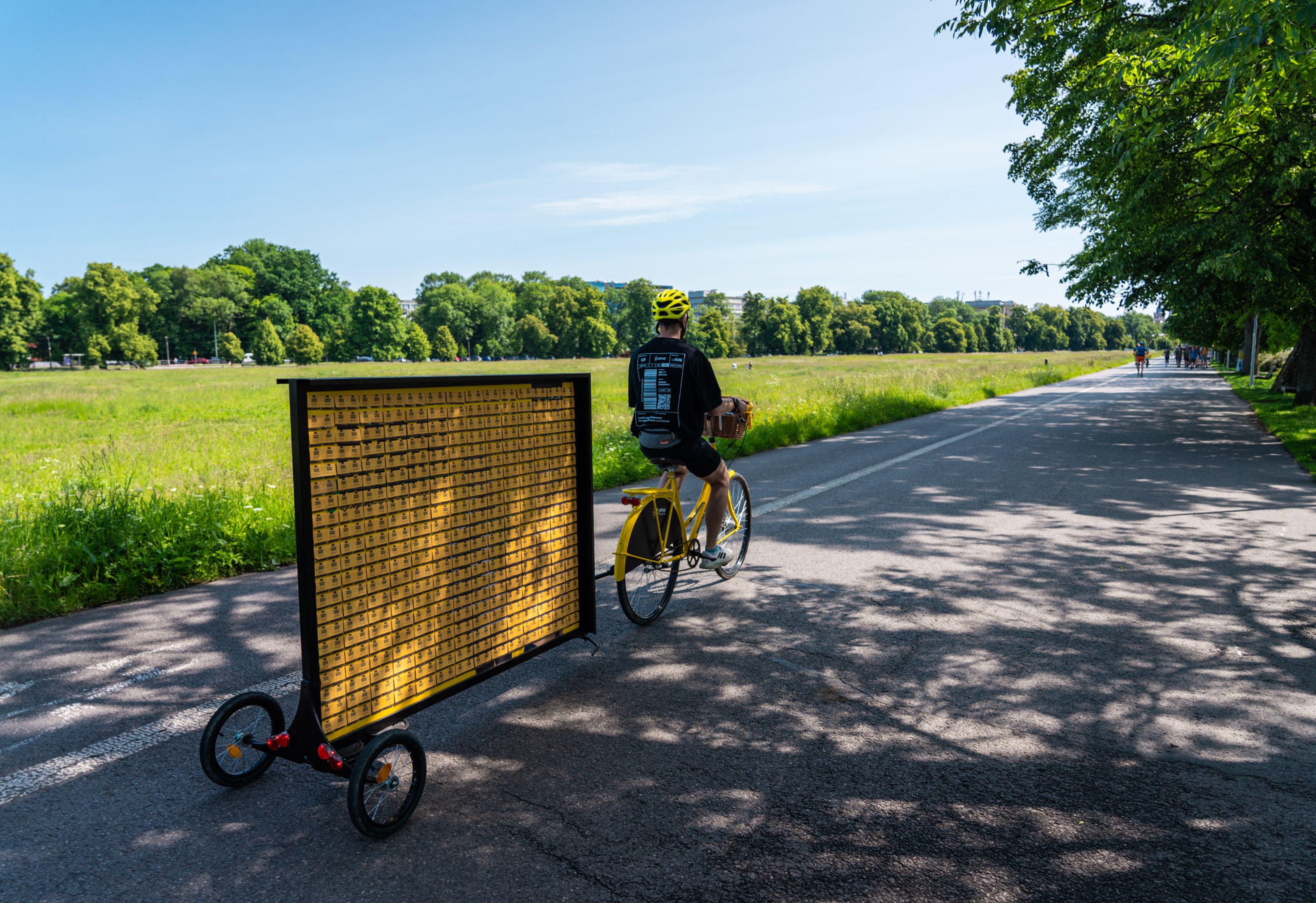 Żółty rower ciągnący tablicę z nasionkami łąki kwietnej, jadący ulicą na tle pola.