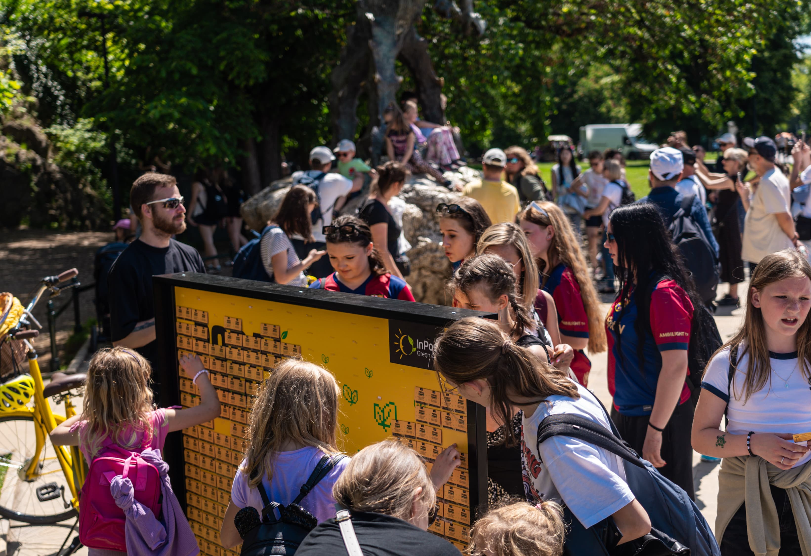 Grupa ludzi w różnym wieku zrywająca opakowania z nasionkami łąki kwietnej z żółtej tablicy.